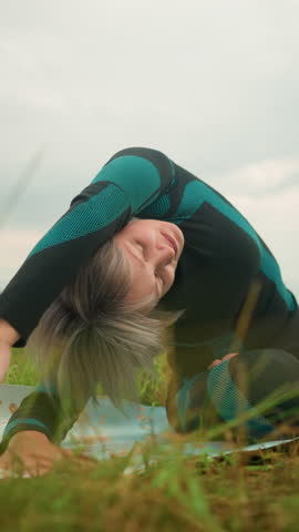 Woman with grey hair lying one sided on yoga mat practicing side bend pose, arms extended, eyes closed, rise up slowly, under cloudy skies in a serene grassy field with trees in the distance
