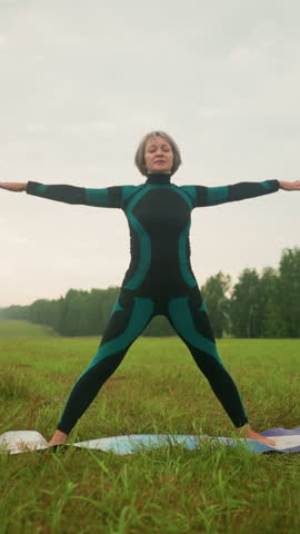 Woman in green and black suit on yoga mat practicing triangle forward pose in serene grassy field under cloudy skies with trees in the background, arms stretched wide