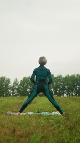 Back view of woman in green-black suit standing on yoga mat practicing triangle pose with arms wide open and legs apart, set against cloudy skies in a peaceful grassy field surrounded by nature