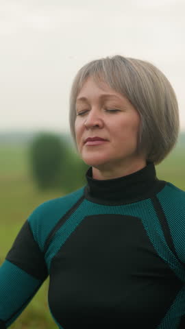 Middle-aged woman in green and black suit practicing yoga outdoors in cow face pose, with eyes closed, focusing on mindfulness and breathing in a serene grassy field under a cloudy sky