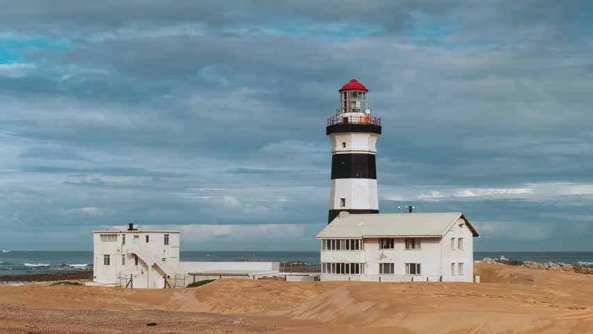 Black and white striped lighthouse overlooks ocean from sandy beach, with small building nearby. Sky is filled with soft clouds. Timelapse of Cape Recife lighthouse in South Africa at dawn