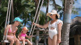 On a vibrant tropical beach, a young mother in a white swimsuit and straw hat joyfully swings her baby and older daughter on a swing, surrounded by palm trees and white sand, slow motion - Powered by Shutterstock - Get 15% off with code: PIKWIZARD15