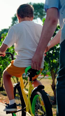 Father teaches kid son to ride bike in park. Child cyclist with Father, nature. Happy family, little boy together with her dad learns to ride bicycle outdoors in summer. Family weekend. Kid