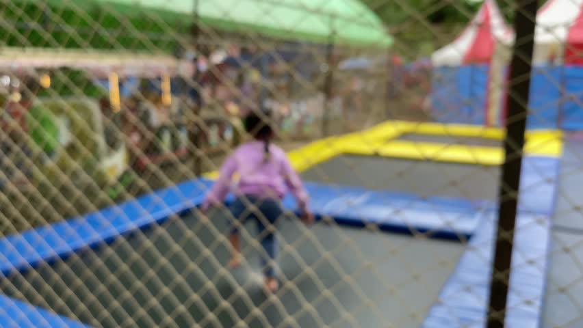 A blurry child in a purple shirt plays on a trampoline with blue and yellow padding, enclosed by a net. The outdoor setting features colorful tents and kiddie rides, creating a festive atmosphere
