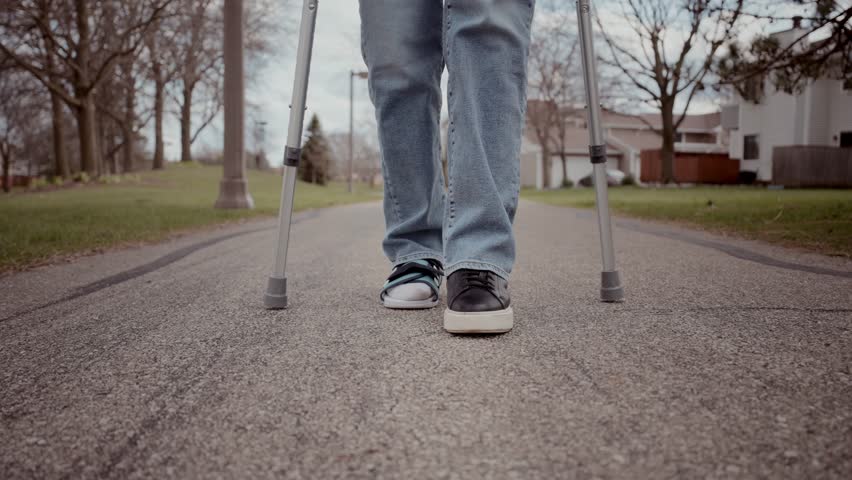Close-up shot of a woman's legs as she walks with crutches, wearing a medical orthopedic walking boot provided in the emergency room. Footage at 60 fps
