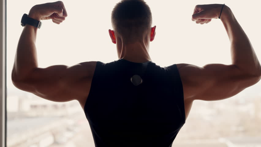 Backlit silhouette of a muscular man flexing his arms, showcasing strength and fitness against a well-lit window backdrop. Strong Athletic Man Flexing Muscles in Silhouette Against Window