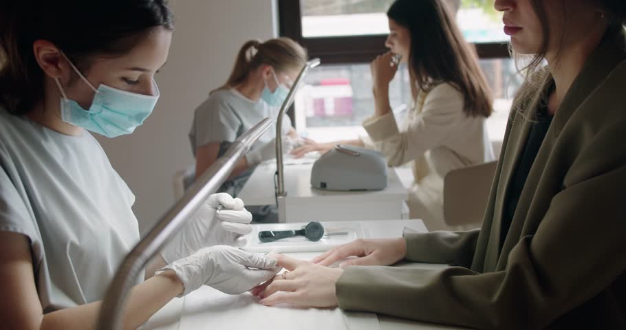Manicurist in mask and gloves treats cuticle with pusher at beauty salon. Hand care procedures. Manicure in a beauty salon. Nail extension. Woman performs medical manicure using special tools.