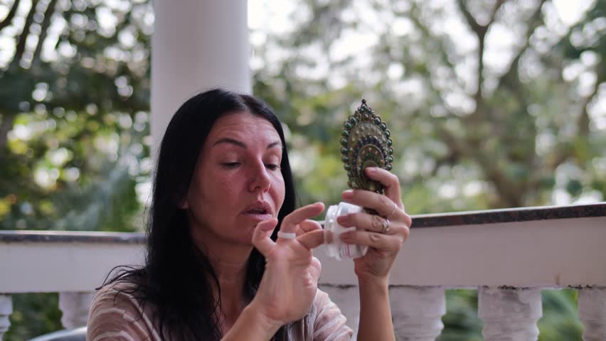 A mature woman gently applies face cream while looking in a hand mirror on a peaceful outdoor veranda.