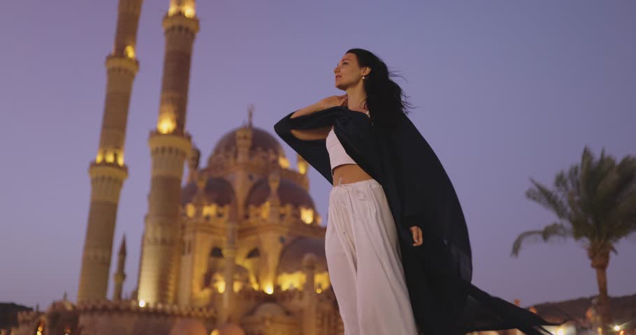 Elegant woman in flowing attire posing in front of the illuminated Sahaba Mosque in Sharm El Sheikh at dusk. Perfect for travel, culture, and lifestyle promotions emphasizing beauty and heritage.