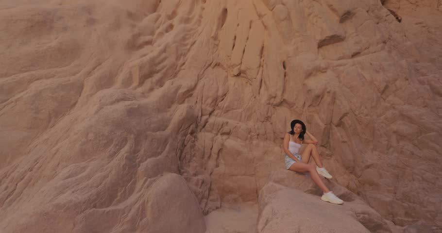An epic wide shot captures a woman relaxing on rocky terrain in Wishwashi Canyon, Egypt, with a dramatic tilt-up view of the towering cliffs. Perfect for travel ads, adventure content, and nature
