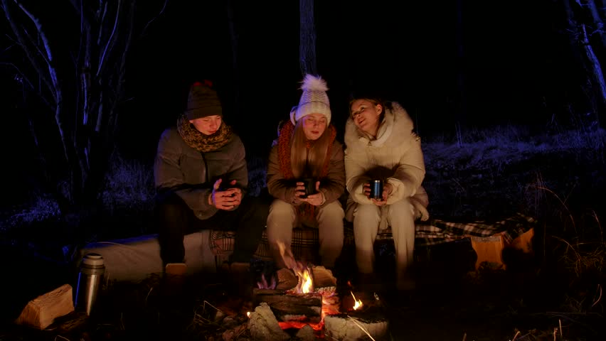 A group of young friends sits around a glowing campfire at night, dressed in cozy winter clothing. They hold metal mugs and a thermos, sharing warm drinks and enjoying the outdoors together.