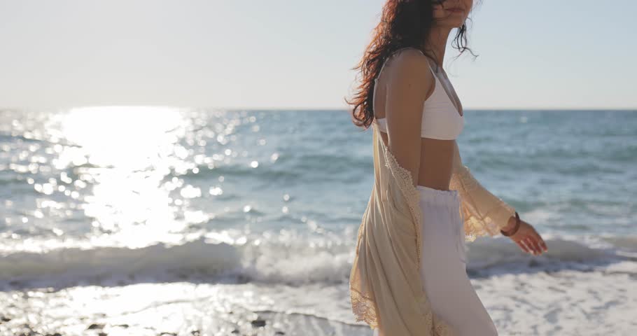Slim, tanned, young Caucasian woman in a stylish boho outfit walking gracefully along the beach in slow motion. Perfect for fashion, lifestyle, and travel-themed visuals. 