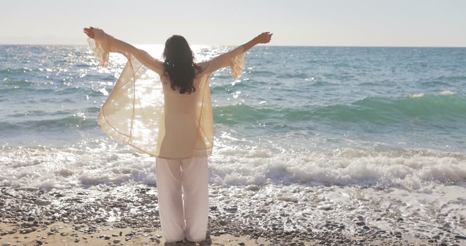 Young woman standing on a rocky beach, arms outstretched to enjoy the ocean breeze and sunlight reflecting on the waves. Perfect for lifestyle, travel, and mindfulness visuals. 
