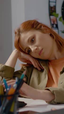 Vertical Screen: Woman types on her computer with a bored expression in her home office. She appears stressed and exhausted by her freelance work at this late hour.