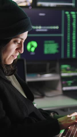Vertical HD video of professional female hacker typing virus code on laptop for cyber attack in dark room with multiple monitors on the background