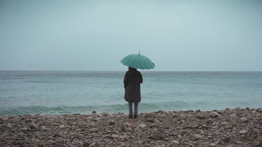 A young woman in a raincoat and carrying an umbrella walks along the beach during the rain