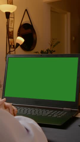 Vertical Screen: Professional businesswoman working on laptop with green screen during video conference, sitting in cozy home office environment during evening hours