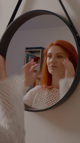 Vertical Screen: Young woman with red hair looking at her reflection in a round mirror, trying different hairstyles and feeling dissatisfied