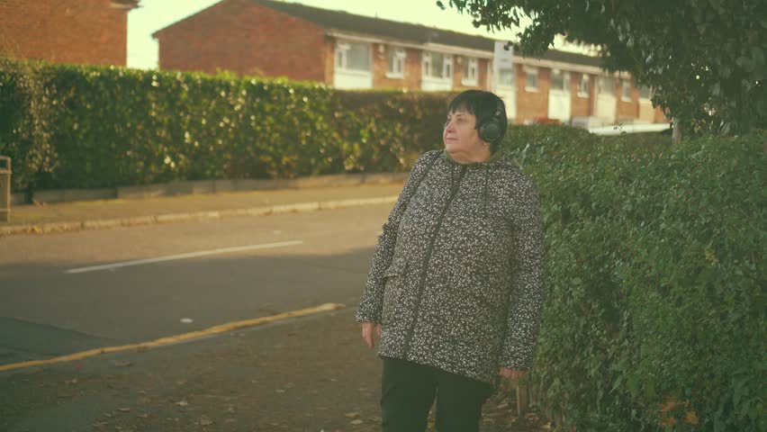 Woman standing on sidewalk wearing headphones. Old female on street enjoying music in earphones. Senior lady on pavement relaxing with audio. Middle-aged woman outdoors listening to tunes