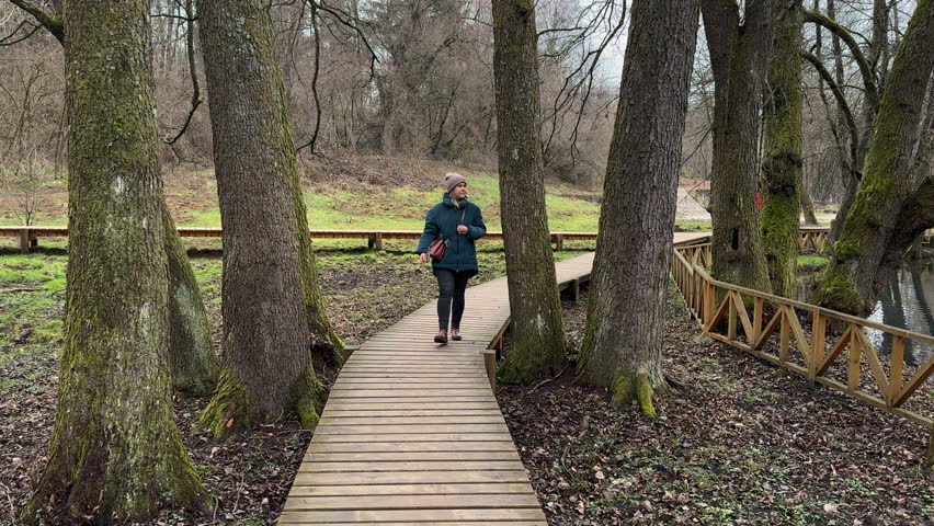 Beautiful woman in blue jacket walking on a wooden pathway surrounded by big trees in a park. Slow motion, winter, daytime, overcast.