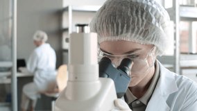 Close up of young Caucasian woman wearing disposable medical hat, goggles and overall doing research using microscope in lab then looking at camera and smiling - Powered by Shutterstock - Get 15% off with code: PIKWIZARD15