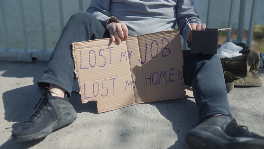 A homeless man sits on a concrete walkway of a bridge, holding a cardboard sign that reads Lost my job, lost my home, revealing the harsh reality of financial struggle and homelessness.