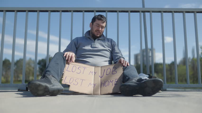 A homeless man sits on a concrete bridge walkway, holding a cardboard sign declaring Lost my job, lost my home.