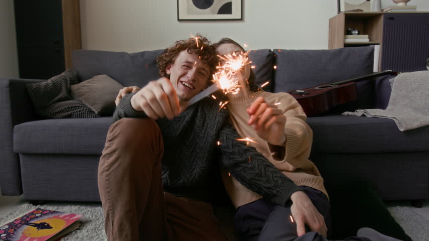 Portrait shot of smiling joyful young couple holds sparklers, leaning against couch in floor of their cozy living room
