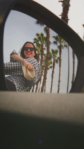Vertical: Excited woman enjoys the palm trees of seaside road. A girl leans out of window of rental car on a coastal highway on a sunny day. Hair in wind. Tropical vacation on beach of exotic islands