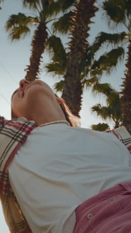 Vertical: Excited woman enjoys the palm trees of seaside road. Girl leans out of window of a rental car on a coastal highway on a sunny day. Hair in wind. Tropical vacation on beach of exotic islands