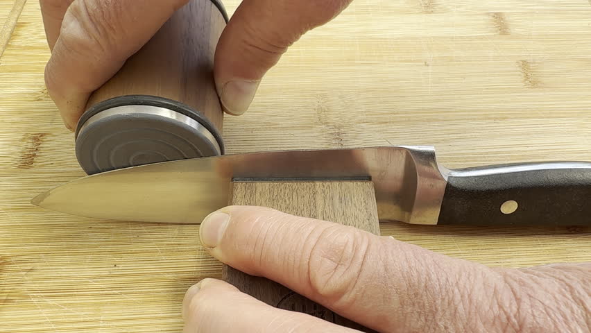 Closeup of hands using a modern knife sharpener, consisting of a magnetic wooden block to hold the knife, while a disc is rolled along the blade length to sharpen it.