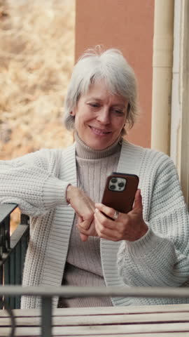Vertical video, Smiling elderly woman enjoys her morning coffee while using her phone on the balcony overlooking the city.