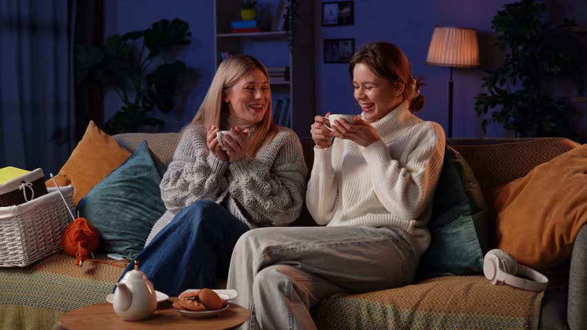 Two women relax on a comfy couch, sharing laughter while knitting with colorful yarn and enjoying warm drinks. The soft glow of the evening creates a cozy atmosphere for creativity.
