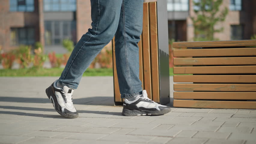 leg view of boy walking toward wooden bench to sit and rest during sunny day in peaceful outdoor setting with visible plants, trash bin, modern buildings, and green lawn in background