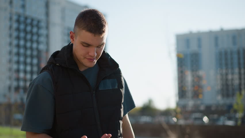 young man in dark vest inhales vape and coughs while holding chest with visible discomfort standing outdoors in urban area with blurred residential high-rise buildings in background