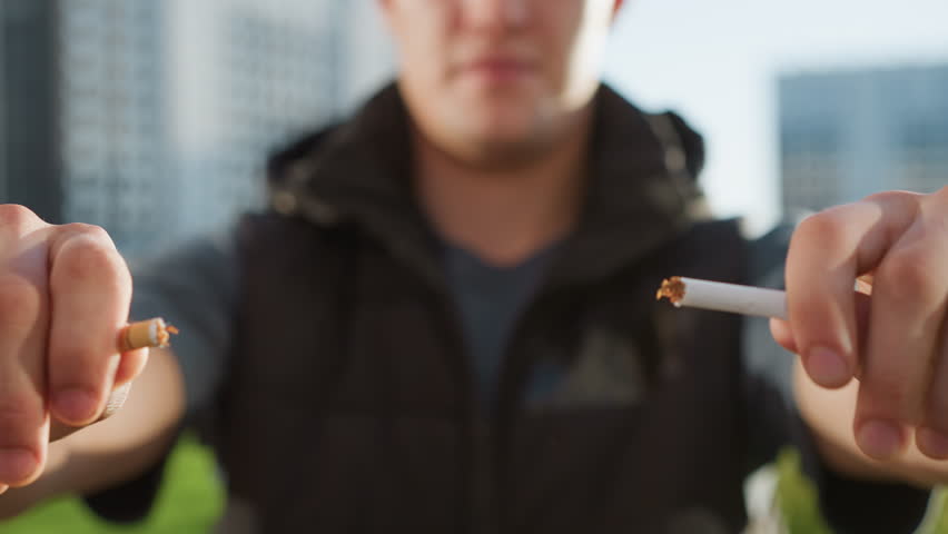 man standing with sunlit hands brought forward gripping and snapping cigarette mid fail gesture in front of blurred building and lush greenery, attempt to quit smoking