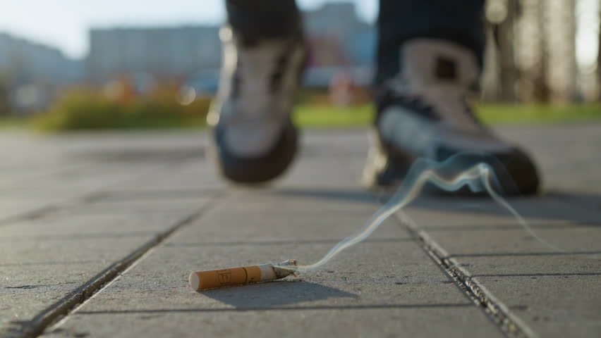 close up of burning cigarette lying on paved sidewalk with smoke swirling upward as person walks toward it and prepares to crush it underfoot