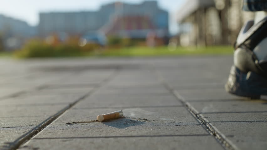close up of burning cigarette lying on paved sidewalk with smoke swirling upward as person walks toward it and prepares to crush it underfoot