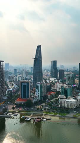 Vertical drone shot of the modern skyline of Ho Chi Minh City featuring the Bitexco Financial Tower, viewed across the Saigon River on a hazy afternoon. Vietnam, UHD.