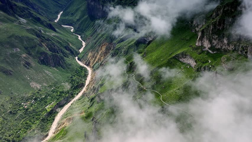 A dramatic drone shot revealing a winding trekking path at high altitude in the Colca Valley, with the misty clouds parting to unveil the rich landscape below during the rainy season.