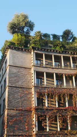 Urban building with lush rooftop garden and facade covered in vibrant climbing plants with red and green leaves, blending architecture with nature, Milan, Italy. Eco friendly urban environment