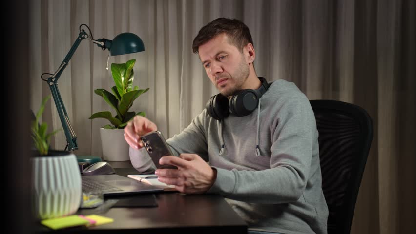Tired freelancer procrastinating at home office, scrolling phone instead of working. Unproductive man using smartphone at home desk. Concept of procrastination and distraction in remote work