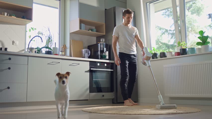 House cleaning with vacuum cleaner and small dog at home. Man vacuuming kitchen floor while curious jack russell terrier dog observing. Pet prevents from doing housework