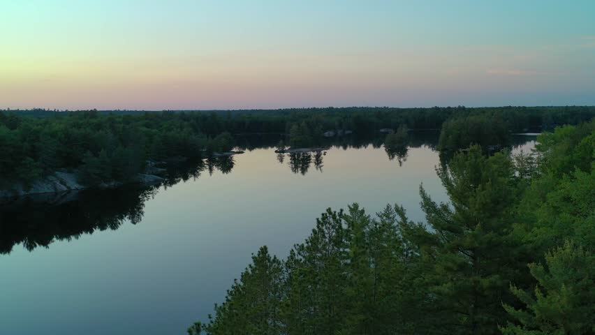 Flying over trees in a forest and a calm reflecting lake at sunset. A cottage with a dock and a boat is on the shoreline during the summer.