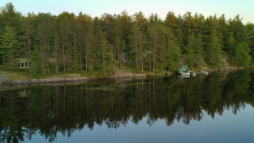 Flying towards a cottage with a dock and boats on the shoreline of a calm lake, reflecting the trees and the sunset during the summer.