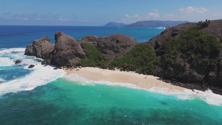 Aerial view of a rugged coastline in Lombok, Indonesia, where turquoise waves crash against dark cliffs. The deep blue ocean contrasts with the greenery on the rocky landmass