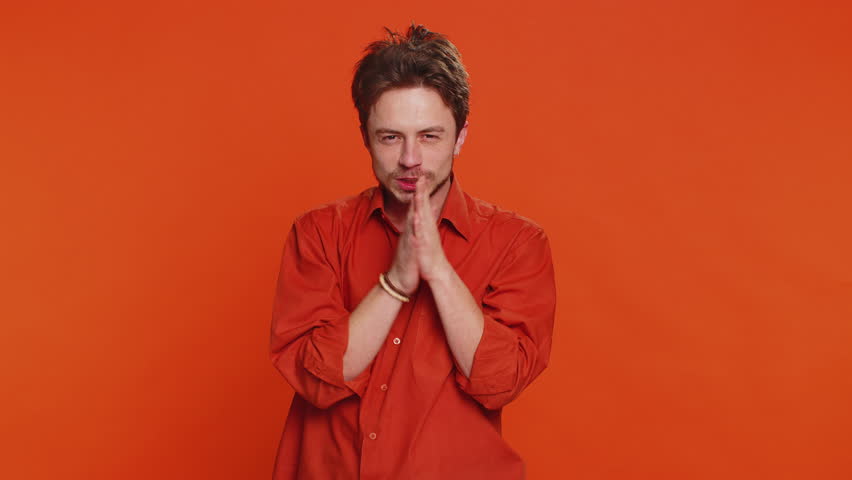 Sneaky cunning Caucasian young man with tricky face gesticulating and scheming evil plan, thinking over devious villain idea, cheats, jokes, pranks. Handsome guy isolated on orange studio background