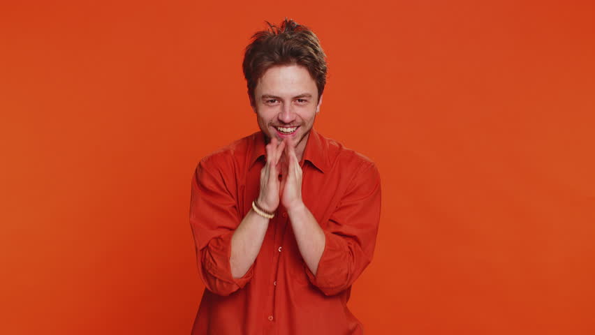 Sneaky cunning Caucasian young man with tricky face gesticulating and scheming evil plan, thinking over devious villain idea, cheats, jokes, pranks. Handsome guy isolated on orange studio background