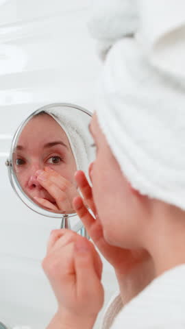 Mature middle-aged woman carefully examining her face in a magnifying mirror, gently touching problem areas with fingers. Thoughtfully assessing skin condition, choosing suitable cosmetic morning care