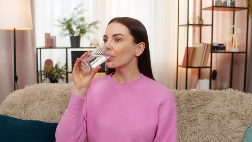 Thirsty young woman at home drinking pure filtered water feeling thirsty after hot weather. Caucasian girl on sofa promoting healthy lifestyle, hydration good habit, satisfied and refreshed emotional